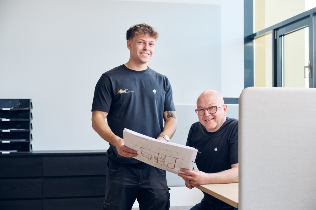 Zwei Männer in Arbeitskleidung besprechen Baupläne in einem Büro mit Fenster