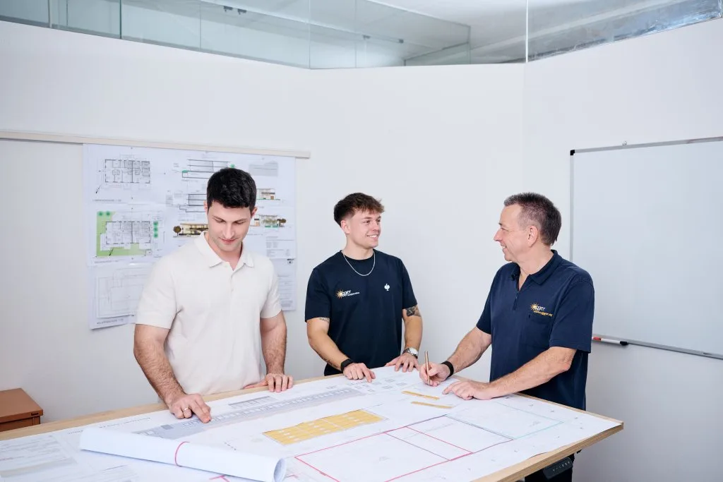 Drei Männer besprechen Baupläne in einem hellen Büro mit Zeichnungen an der Wand