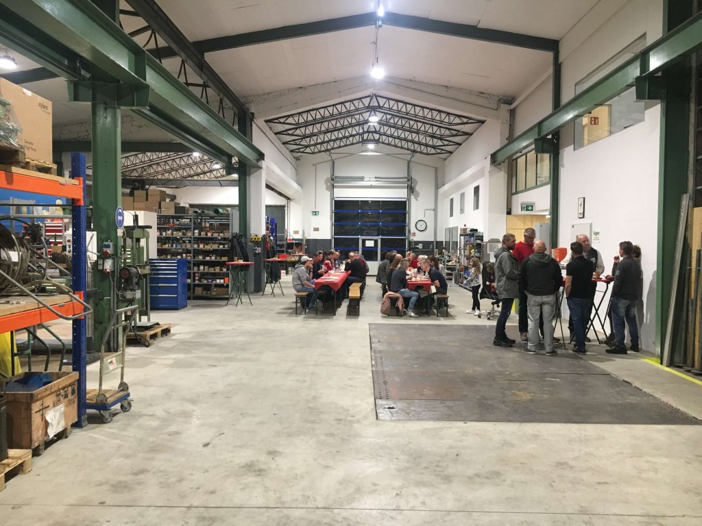 Blick in eine große Werkhalle mit Menschen, die an Tischen sitzen und stehen